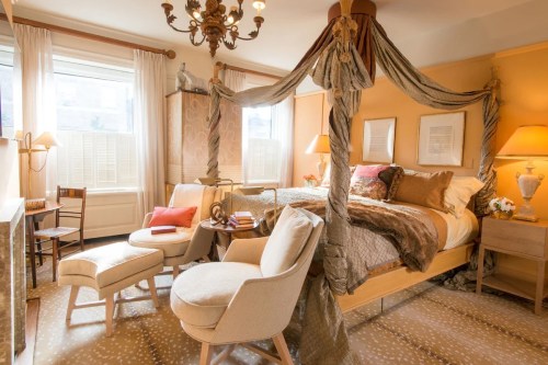 Cozy bedroom with a four-poster bed, soft lighting, and elegant decor, featuring chairs and a warm color palette.