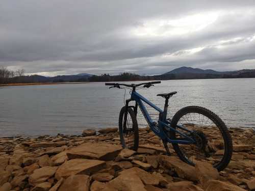 A blue mountain bike rests on rocky shore by a calm lake, with mountains and cloudy skies in the background.