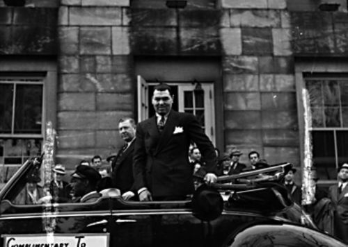 A man in a suit stands in a convertible car, with a crowd and historic building in the background.