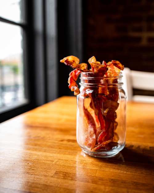 A glass jar filled with crispy bacon strips, placed on a wooden table near a window.