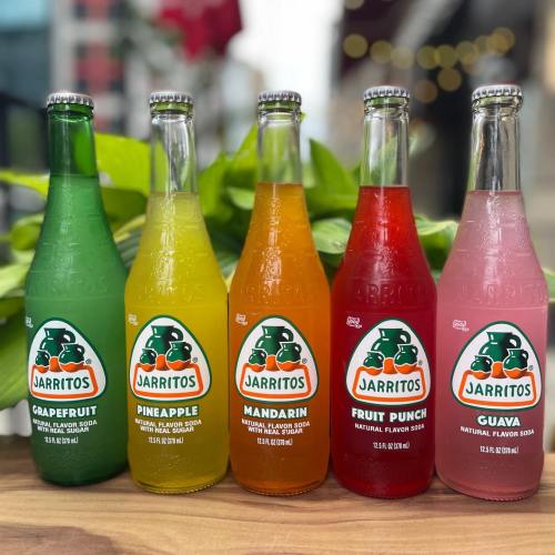 Five colorful bottles of Jarritos soda: grapefruit, pineapple, mandarin, fruit punch, and guava, arranged on a table.