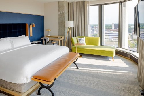 A modern hotel room featuring a large bed, a green couch, and a view through large windows.