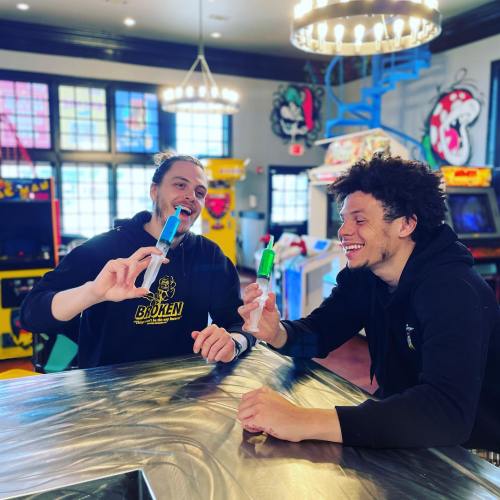 Two friends smile and hold colorful popsicles while sitting at a table in a vibrant arcade setting.