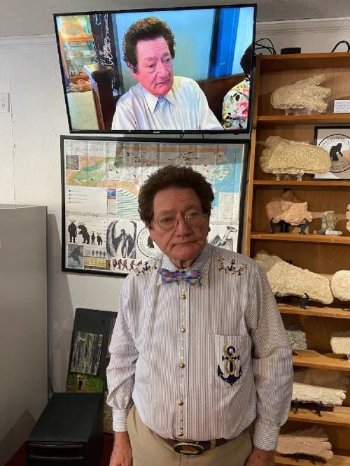 A man in a striped shirt and bow tie stands in front of a display with a TV showing his image.