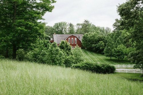 A red barn-style house surrounded by lush green trees and a grassy field under a cloudy sky.