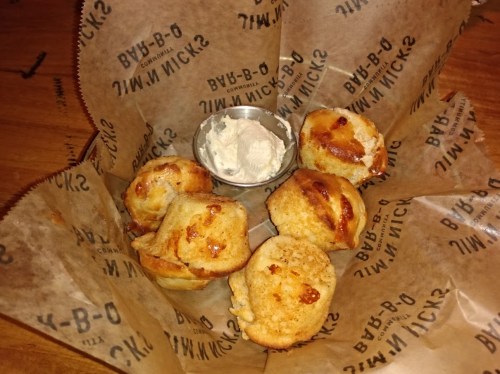 A basket of golden-brown muffins with a small cup of creamy dip, all lined with branded paper.