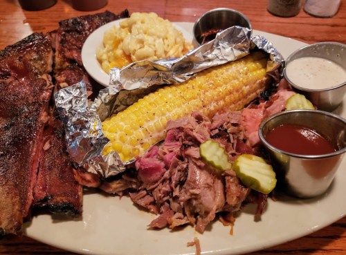 A plate of barbecue featuring ribs, pulled pork, corn on the cob, macaroni and cheese, and dipping sauces.
