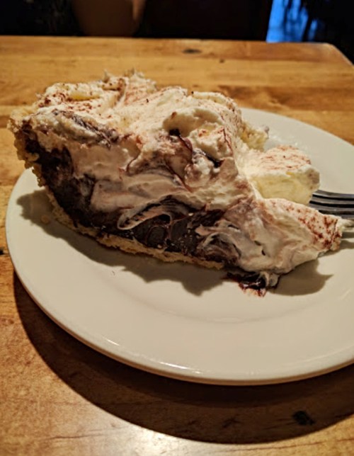 A slice of chocolate cream pie topped with whipped cream on a white plate, resting on a wooden table.