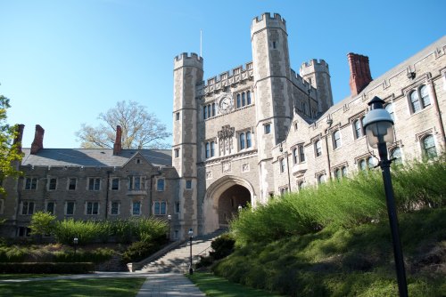A historic stone building with towers, surrounded by greenery and a clear blue sky. Pathway leads to the entrance.
