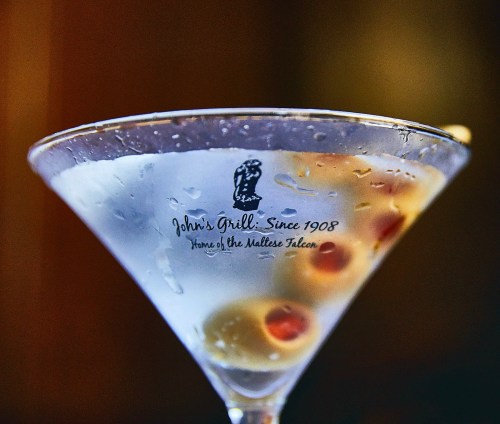 A close-up of a martini glass with olives, featuring the logo of John's Grill, established in 1908.