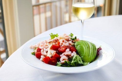 A fresh salad with shrimp, cherry tomatoes, and avocado, accompanied by a glass of white wine on a table.