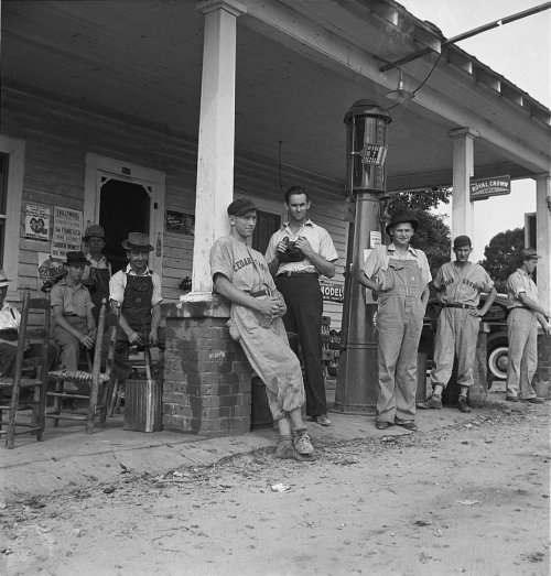 A group of men in work clothes gathered outside a gas station, some sitting and others standing, in a rural setting.