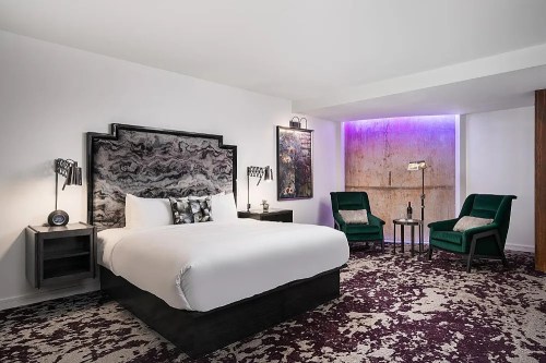 Modern hotel room featuring a king bed, stylish decor, two green chairs, and ambient lighting.
