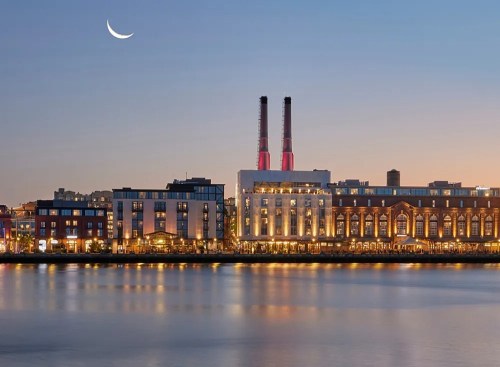 A serene waterfront view at dusk, featuring buildings and smokestacks, with a crescent moon in the sky.