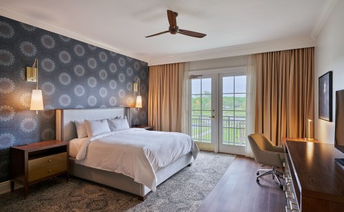 A modern hotel room featuring a queen bed, patterned wallpaper, a desk, and large windows with curtains.