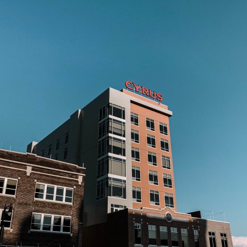A tall building with a red "CYRUS" sign against a clear blue sky, next to a brick structure.