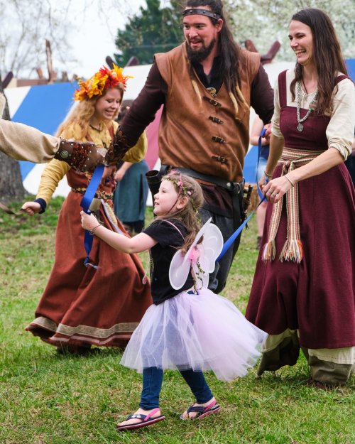 A group of people in medieval costumes joyfully dance, with a young girl in a fairy outfit leading the way.