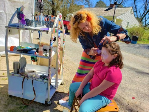 A woman applies face paint to a young girl sitting on a chair, with a colorful cart of supplies nearby.
