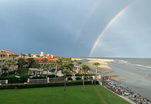 A scenic view of a beach with a rainbow, palm trees, and a resort under a cloudy sky.
