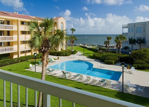A sunny beachfront view featuring a pool surrounded by palm trees and green lawns, with ocean waves in the background.