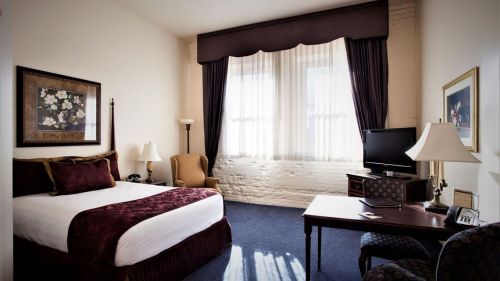 Cozy hotel room with a queen bed, armchair, desk, TV, and large windows with sheer curtains.