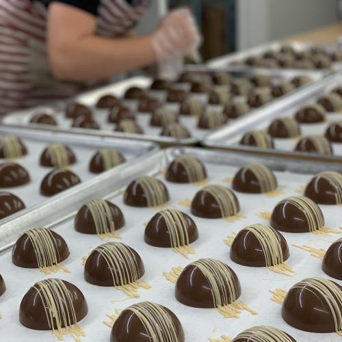 Trays of chocolate confections drizzled with white chocolate, with a person decorating in the background.