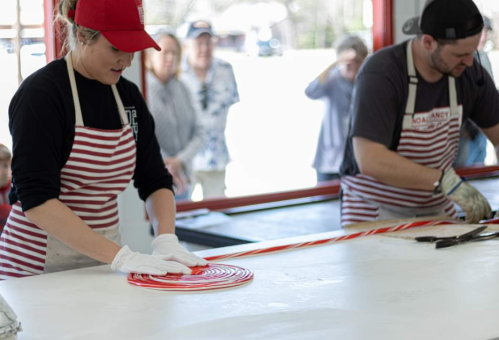 A woman in a red cap and striped apron shapes candy on a table, while a man in a similar outfit assists nearby.