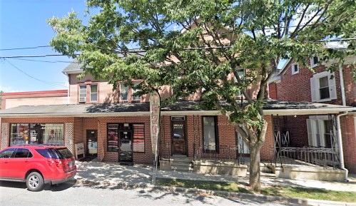 A brick building with shops and a red car parked outside, shaded by a large tree on a sunny day.