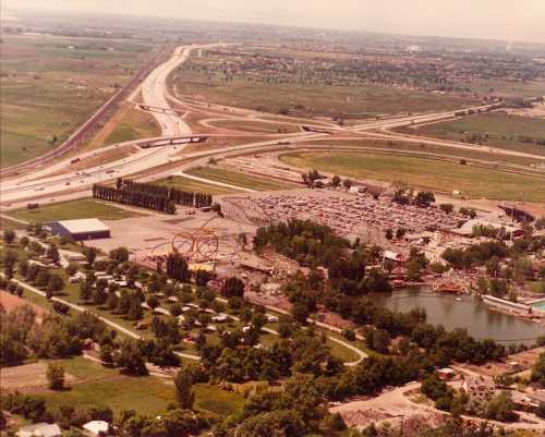 Aerial view of a highway, amusement park, and parking lot surrounded by greenery and open fields.