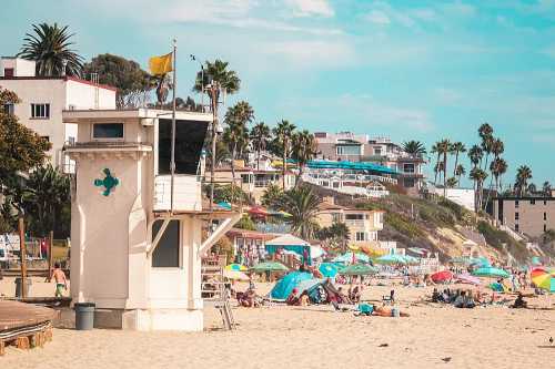 A beach scene featuring a lifeguard tower, colorful umbrellas, and people enjoying the sun with palm trees in the background.