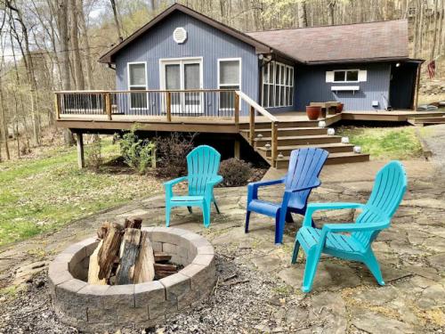 A cozy blue house with a deck, surrounded by trees, featuring a fire pit and colorful chairs in the foreground.