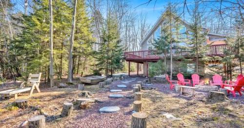 A cozy cabin in the woods with a deck, surrounded by trees, a fire pit, and seating areas on a gravel path.