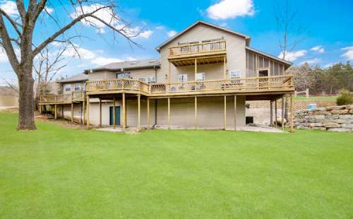 A large wooden deck surrounds a two-story house, set on a green lawn with trees and a clear blue sky in the background.