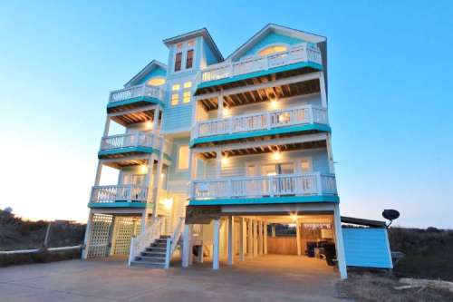 A large, multi-story beach house with balconies, illuminated at dusk, surrounded by sandy terrain.