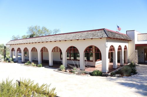 A single-story building with arched windows and a red roof, surrounded by desert landscaping and a clear blue sky.