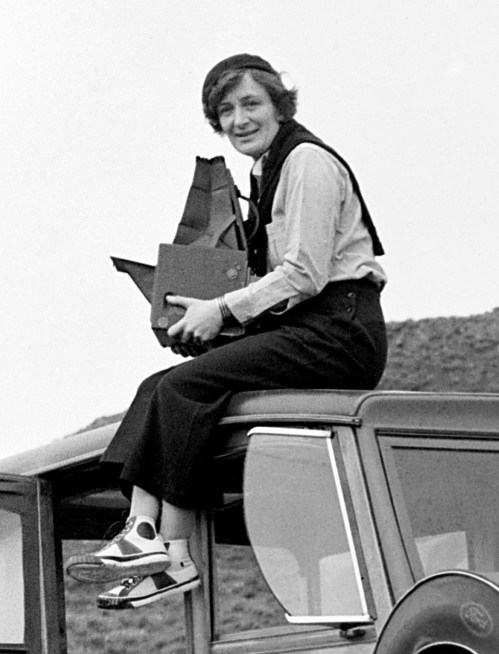 A woman sits on the roof of a vintage car, holding a camera and smiling, dressed in a blouse and trousers.