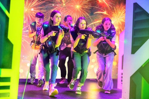 A group of five kids in tactical vests, holding laser guns, moving cautiously against a colorful fireworks backdrop.
