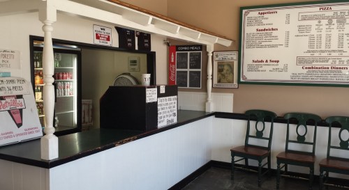 A small restaurant counter with a menu board, drinks fridge, and empty chairs in a casual dining area.