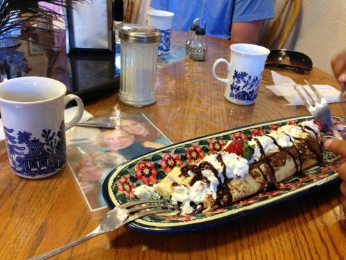 A colorful plate of dessert crepes drizzled with chocolate and topped with whipped cream, coffee cups in the background.