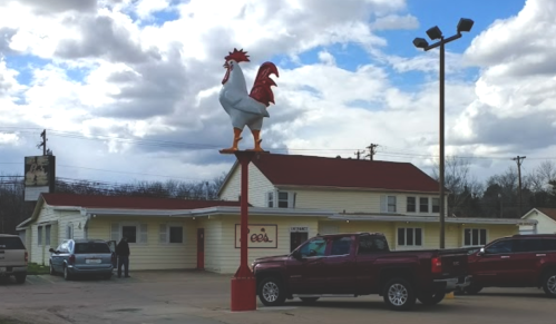 A large rooster statue atop a pole outside a building, with cars parked nearby under a cloudy sky.