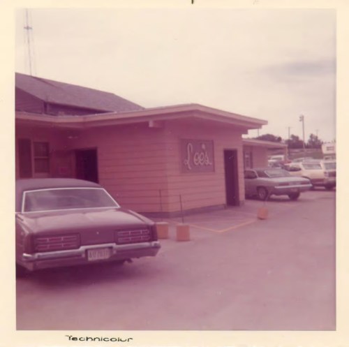 A vintage photo of Lee's building with two parked cars in the foreground and a cloudy sky above.