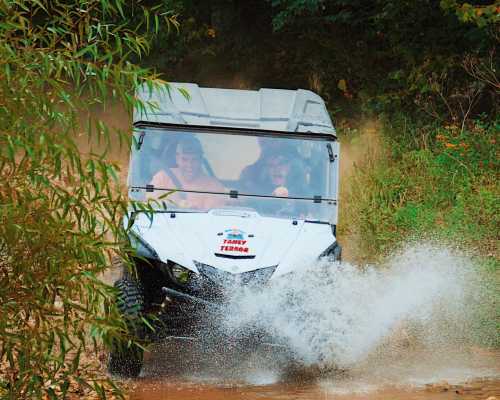 A white ATV splashes through muddy water, with two excited passengers enjoying the ride amidst greenery.