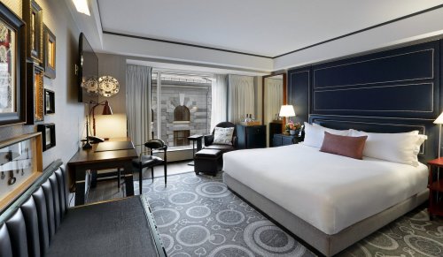 A modern hotel room featuring a queen bed, desk, armchair, and large window with city views. Elegant decor throughout.