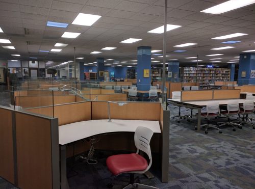 A spacious, empty library with rows of study desks and chairs, illuminated by overhead lights.