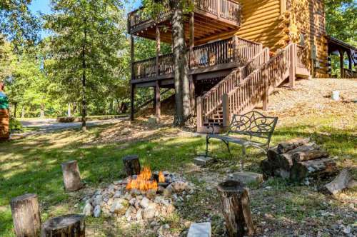 A cozy outdoor scene featuring a fire pit surrounded by logs, with a cabin and trees in the background.