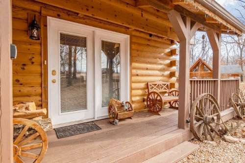 A cozy log cabin porch with double doors, wooden benches, and rustic wagon wheels.