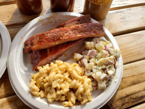 A plate with two pieces of smoked ribs, creamy macaroni and cheese, and a potato salad on a wooden table.