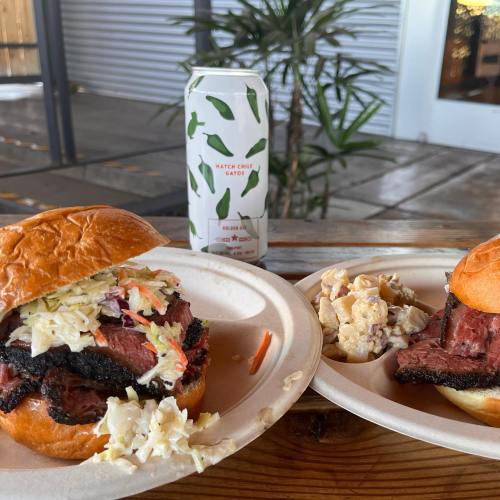 A close-up of a smoked meat sandwich with coleslaw, alongside a side of potato salad and a can of hatch chili soda.