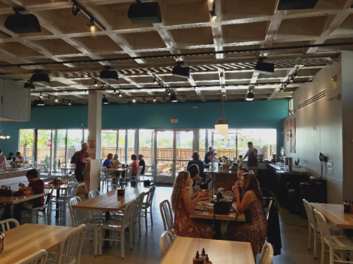 Bright restaurant interior with wooden tables, patrons dining, and large windows letting in natural light.