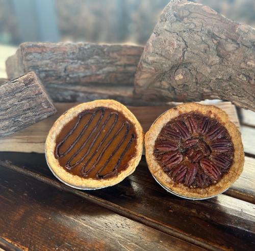 Two pies on a wooden surface: one with chocolate drizzle and the other topped with pecans, surrounded by logs.